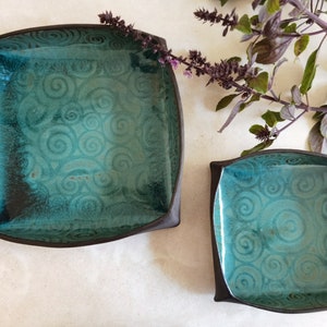 Könnte beinhalten: Zwei quadratische Keramikschalen mit einer teal-farbenen Glasur und einem wirbelnden Muster. Die Schalen befinden sich auf einer weißen Oberfläche.