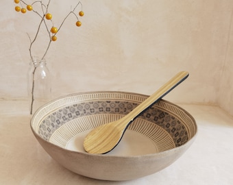 May include: A beige ceramic bowl with a decorative patterned rim and a wooden spoon. The bowl has a wide, shallow shape, and the spoon rests inside. A clear glass vase with a branch of yellow berries is in the background.