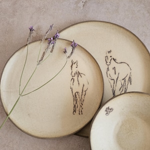Può includere: Un set di piatti in ceramica beige con un'illustrazione di cavallo disegnata a mano in marrone scuro. I piatti hanno un'estetica rustica e terrosa, con un design semplice ed elegante. Un rametto di fiori viola aggiunge un tocco di colore.