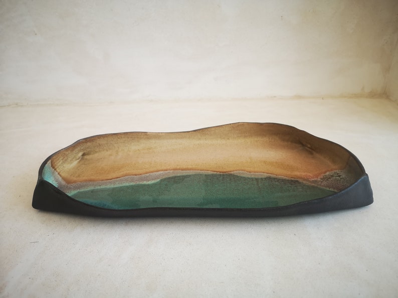 Puede incluir: Una bandeja rectangular de cer&aacute;mica larga con un borde negro y un esmalte verde y marr&oacute;n. La bandeja tiene un borde ligeramente ondulado y una forma org&aacute;nica &uacute;nica.