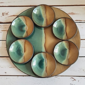 May include: A round ceramic serving tray with six smaller bowls. The tray and bowls are decorated with a green and brown glaze, creating a marbled effect.
