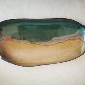 Puede incluir: Un plato de servir de cer&aacute;mica con un esmalte verde, marr&oacute;n y beige. El plato tiene forma de &oacute;valo largo y un borde ligeramente irregular.