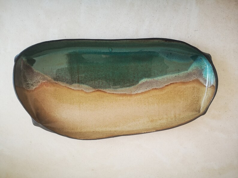 Puede incluir: Un plato de servir de cer&aacute;mica con un esmalte verde, marr&oacute;n y beige. El plato tiene forma de &oacute;valo largo y un borde ligeramente irregular.