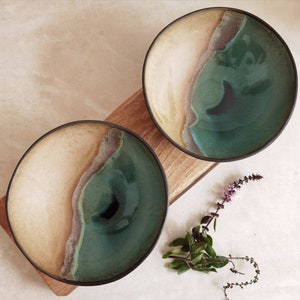 May include: Two ceramic bowls with a green and brown glaze. The bowls have a unique design that resembles a beach scene with waves and sand.