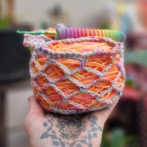 May include: A crocheted yarn bowl with a rainbow coloured yarn inside. The bowl is made of a beige crochet thread and has a textured pattern. The yarn is a mix of orange, yellow, and pink.