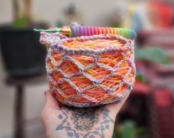May include: A crocheted yarn bowl with a rainbow colored yarn inside. The bowl is made of a beige crochet thread and has a textured pattern. The yarn is a mix of orange, yellow, and pink.