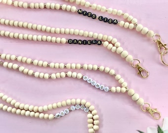 Puede incluir: Cuatro cordones hechos de cuentas de madera natural de color claro intercaladas con pequeñas cuentas metálicas doradas. Cada cordón presenta una sección personalizada con cuentas de letras que deletrean MISS LISA, DANIELA, RACHEL y MRS LOWE, terminando en cierres de metal dorado.