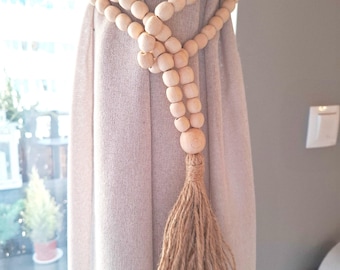 May include: A wooden bead curtain tieback with a natural jute tassel. The tieback is wrapped around a beige curtain.