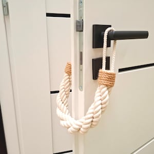 以下が含まれることがあります： 白いロープ製のドアストッパーに茶色のアクセントが施され、黒いドアノブが付いた白いドアに掛けられています。