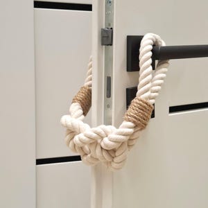 May include: A white rope doorstop with a brown jute accent. The rope is tied in a knot and is hanging on a white door.