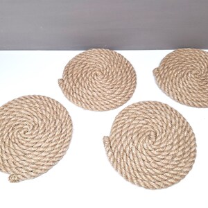 May include: Four round, natural-colored, woven rope coasters. The coasters are made of thick, twisted rope and have a spiral pattern.