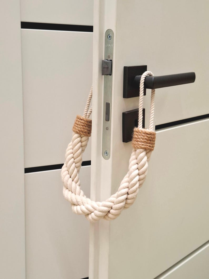 May include: A white rope doorstop with brown ends, hanging on a white door with a black door handle.