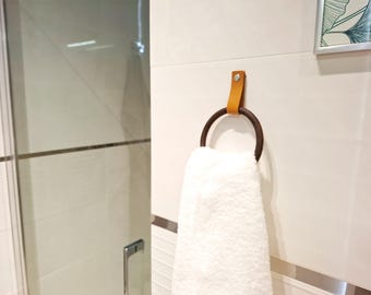 May include: A brown leather towel ring with a white towel hanging on it. The ring is mounted on a white tiled wall.