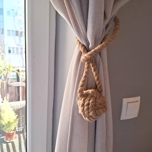 May include: A decorative curtain tieback made of thick, twisted rope. The tieback is knotted into a ball shape and is used to hold back a light-colored curtain. The background includes a window and a wall.