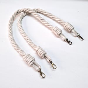 May include: Two white cotton rope handles with brass swivel clasps. The handles are knotted at each end.