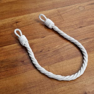 May include: A white rope handle with looped ends, lying on a wooden surface. The rope is tightly woven, and the loops are secured with knots. The handle is likely for a bag or other carrying item.
