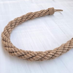 Könnte beinhalten: Ein dicker, naturfarbener Türstopper aus Seil in hellbrauner Farbe. Das Seil ist geflochten und hat an jedem Ende eine Schlaufe.