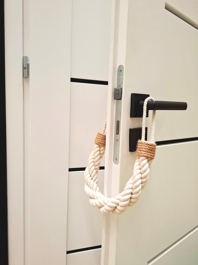 May include: White door with a black door handle and a white rope doorstop. The rope doorstop is tied in a loop and has a brown jute accent.