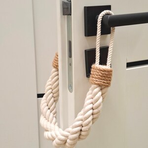 May include: A white rope doorstop with a brown jute wrap. The doorstop is hanging on a white door with a black door handle.
