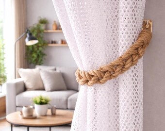 May include: White lace curtains held back by a thick, braided rope tieback. The rope is a natural tan color, and the curtains are hanging in a bright, airy room. The tieback is attached to a small wooden bracket.