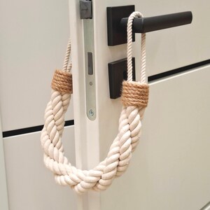 May include: A white rope doorstop with a brown braided rope accent. The doorstop is hanging on a white door with a black door handle.