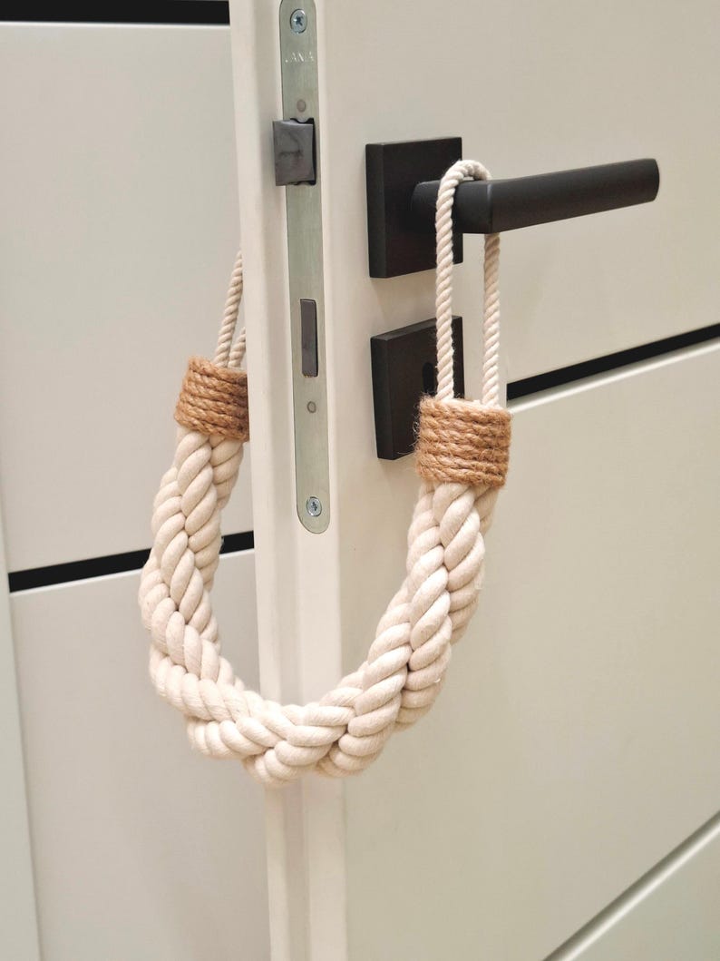 May include: A white rope doorstop with a brown braided rope accent. The doorstop is hanging on a white door with a black door handle.