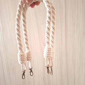 May include: Two white cotton rope handles with metal clasps. The handles are twisted together and have a knot at the end.