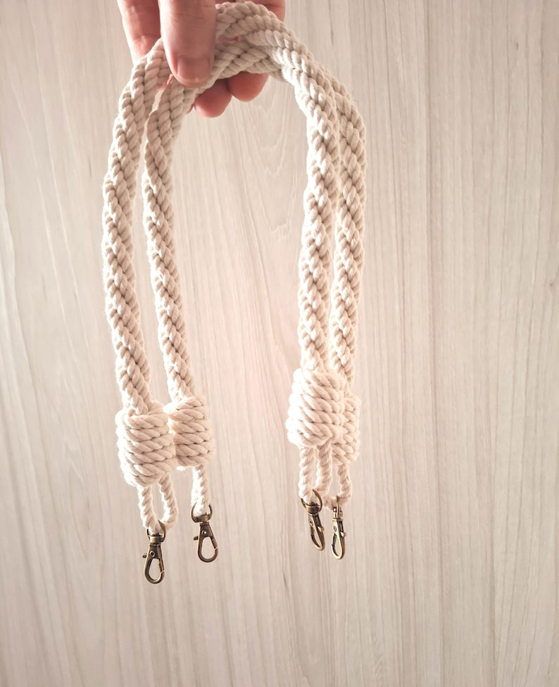 May include: Two white cotton rope handles with metal clasps. The handles are twisted together and have a knot at the end.