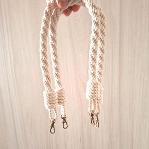 May include: Two cream-colored cotton rope handles with gold metal clasps. The handles are knotted at the top and have a braided design.
