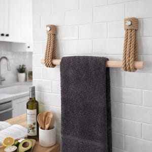 May include: A rustic towel rack made of natural rope and light-colored wood, mounted on a white tiled wall. A dark gray towel hangs from the rack. The rack is in a kitchen setting.