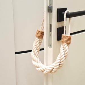 May include: A white rope doorstop with brown rope accents. The doorstop is hanging on a white door with a black door handle.