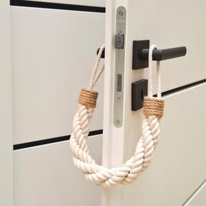 May include: A white rope doorstop with brown jute ends. The doorstop is hanging on a white door with a black door handle.