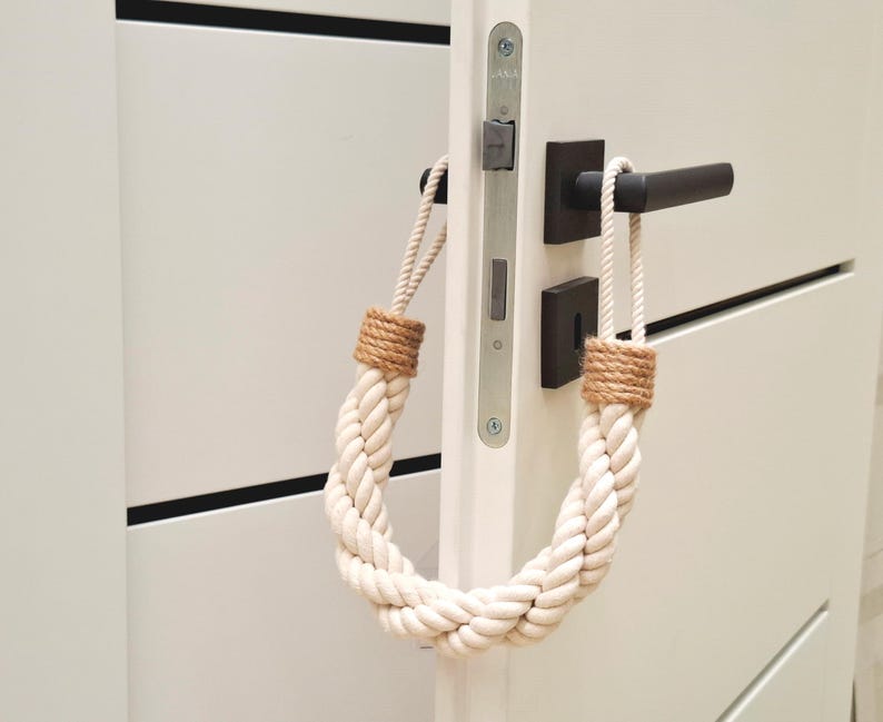 May include: A white rope doorstop with brown jute ends. The doorstop is hanging on a white door with a black door handle.