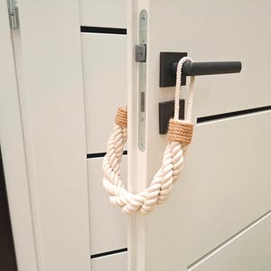 May include: A white door with a black door handle and a rope doorstop. The rope doorstop is made of natural fiber and is in the shape of a loop.