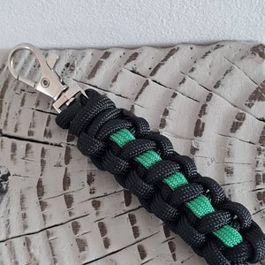 May include: Black and green braided keychain with a silver clasp. The keychain is made of woven black cord with a green stripe. The clasp is a swivel style. The keychain is approximately 15 cm long.