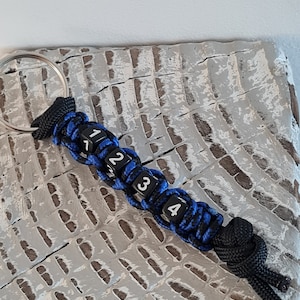 May include: A black and blue paracord keychain with a silver key ring and four black dice with white numbers 1, 2, 3, and 4.
