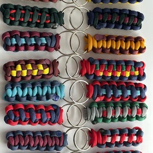 May include: A collection of 12 colourful paracord keychains with different colour combinations. Each keychain features a silver key ring.
