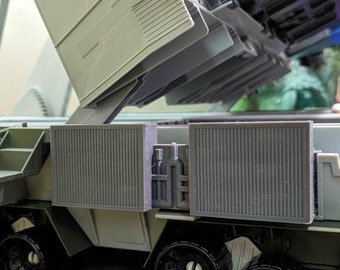 May include: A gray and black toy military vehicle with a raised platform and a large, gray, rectangular cargo container. The vehicle has four large, black wheels and a gray, textured surface.