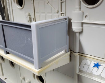 May include: A section of a light gray toy playset with a gray railing and a gray container. The playset has a white base with round openings and the words "G.I. Joe" with a blue star.