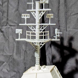May include: A light gray model of a ship's mast, featuring multiple platforms with various antenna-like structures and a ladder. The mast is mounted on a rectangular base, with a detailed railing and support beams. The model is set against a dark gray backdrop.