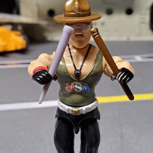 May include: A toy action figure wearing a brown hat, a green tank top with the word "USA" printed on it, and black pants. The figure is holding two sticks in each hand.