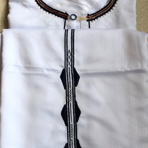 May include: White long-sleeved shirt with a decorative collar and a vertical design on the front. The collar has a dark blue, gold, and red trim. The design on the front is a dark blue and white pattern.