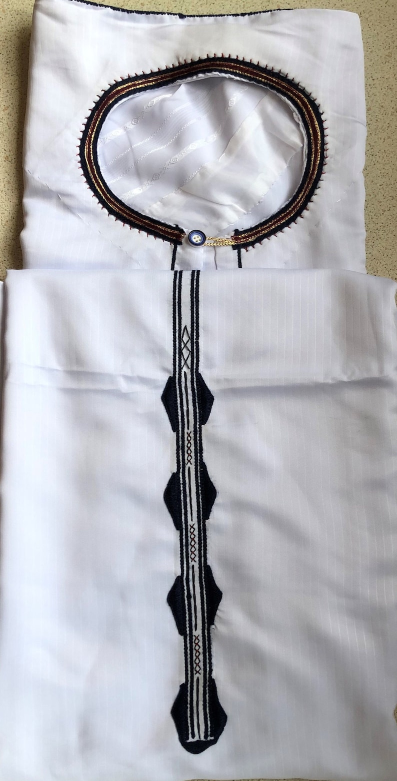 May include: White long-sleeved shirt with a decorative collar and a vertical design on the front. The collar has a dark blue, gold, and red trim. The design on the front is a dark blue and white pattern.