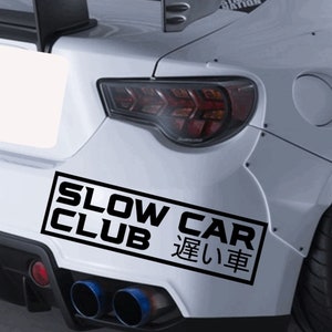 May include: A white car with a black bumper sticker that reads "Slow Car Club" in English and "遅い車" in Japanese.
