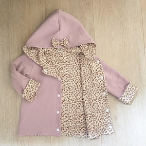 Könnte beinhalten: Hellrosa Kapuzenjacke mit Schleife auf der Kapuze. Die Jacke hat eine Waffelstrick-Außenseite und ein Innenfutter und Manschetten mit Leopardenmuster. Sie wird mit weißen Knöpfen vorne geschlossen.