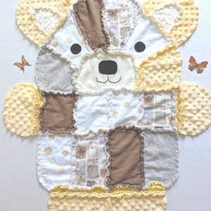 Op de afbeelding: Een handgemaakte patchwork babydeken in de vorm van een beer. De deken heeft verschillende stoffen in crème, bruin en geel. De beer heeft zwarte ogen en een zwarte neus. Decoratieve vlinders zijn rondom de deken geplaatst.