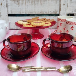 Può includere: Due tazze da tè rosse con piattini e un set di cucchiaini di bambù dorati su una tovaglia a strisce bianche e rosse. Le tazze da tè sono riempite di un liquido scuro. Un piatto di biscotti si trova su un supporto per torte a piedistallo rosso sullo sfondo.