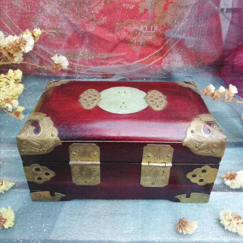 May include: A rectangular, dark red wooden box with brass corner accents and hinges. The box features a central, oval-shaped jade inlay and decorative brass embellishments. The box is closed.