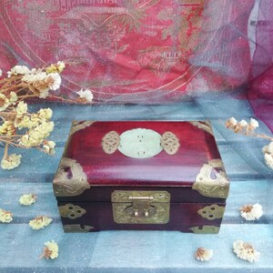 May include: A rectangular, dark red wooden box with brass corner accents and a central clasp. The lid features a light green oval-shaped jade piece with decorative brass elements. The box is closed and sits on a blue surface.