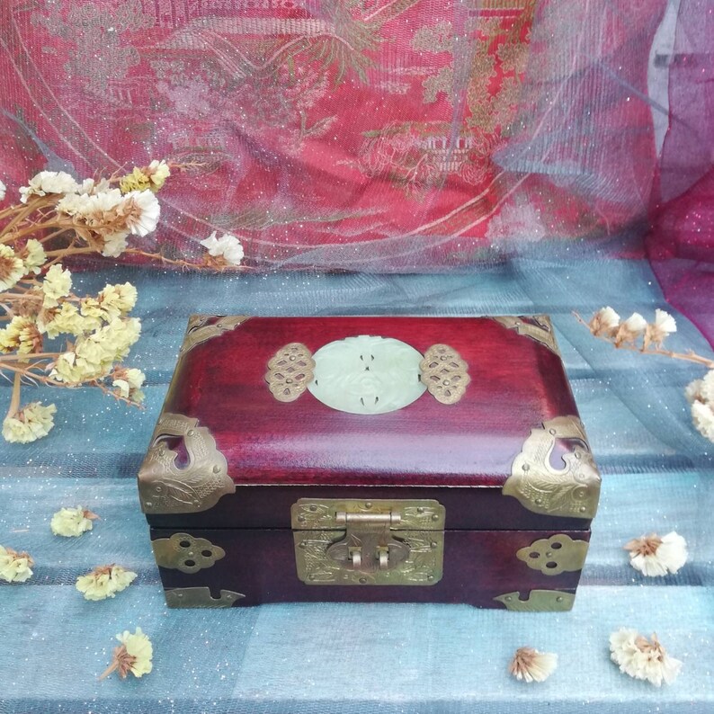 May include: A rectangular, dark red wooden box with brass corner accents and a central clasp. The lid features a light green oval-shaped jade piece with decorative brass elements. The box is closed and sits on a blue surface.